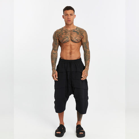 Front view of men’s black paneled drop-crotch capri pants with elastic waistband and relaxed avant-garde silhouette.